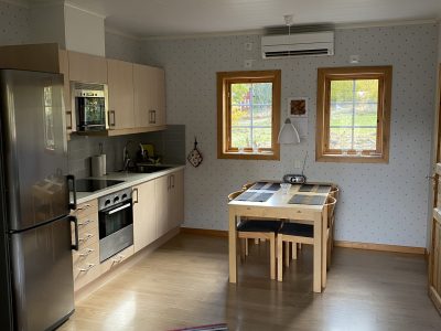 Ett ljust kök med ljusa träskåp, en modern spis och en kyl/frys. En matplats med ett träbord och stolar står vid två fönster. Randiga mattor ligger på trägolvet.