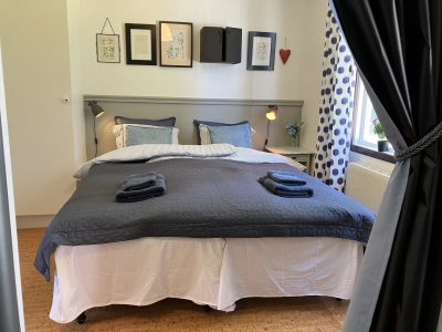 Ett sovrum med en stor dubbelsäng bäddad med ett mörkblått överkast, omgiven av sänglampor och tavelramar på väggen. Ett fönster med mörkblåprickiga gardiner släpper in ljus.