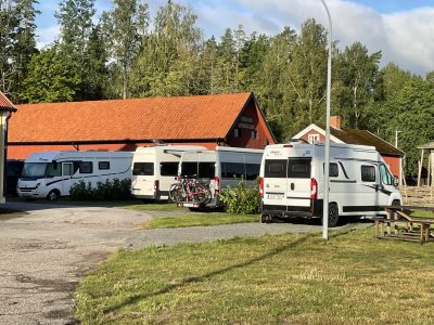 Fyra husbilar står parkerade vid en röd ladugård i lantlig miljö med omgivande grönska och träd.