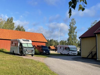 Tre husbilar parkerade på en asfalterad väg mellan röda och gula byggnader på en solig dag.