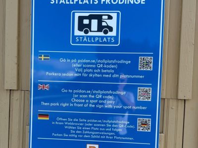 En blå informationsskylt för "Ställplats Frödinge" med instruktioner och en QR-kod på svenska, engelska och tyska.