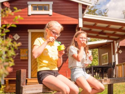Två unga tjejer sitter på ett staket framför en röd stuga och äter glass.