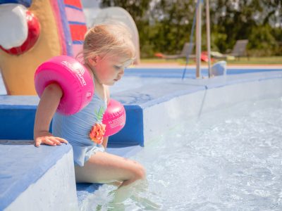 En flicka med rosa armpuffar sitter vid kanten av en pool och plaskar med fötterna.