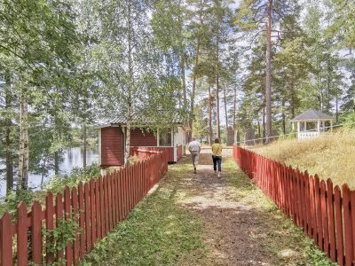 En stig kantad av ett rött trästaket leder mot en röd stuga vid en sjö, med två personer som promenerar bort från betraktaren. I bakgrunden syns skog och en vit pergola.
