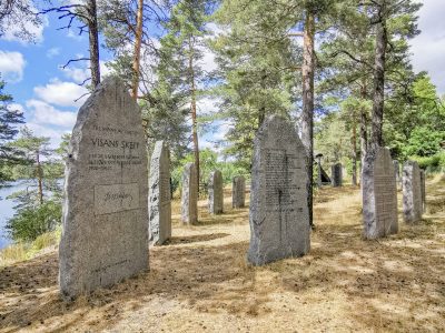 En grupp upprättstående stenar med inskriptioner står på en kulle täckt av gräs och tallbarr. I bakgrunden syns höga träd och en sjö. En av stenarna är märkt "VISANS SKEPP".