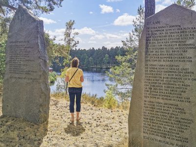 En kvinna i gul tröja och jeans står mellan två stora stenar med inskriptioner, och blickar ut över en lugn sjö omgiven av skog. En av stenarna har en text med rubriken "LYCKAN".