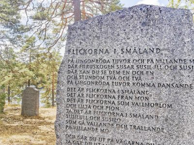 En detaljbild av en stor sten med en inskription med titeln "FLICKORNA I SMÅLAND", omgiven av träd och med en sjö synlig i bakgrunden.