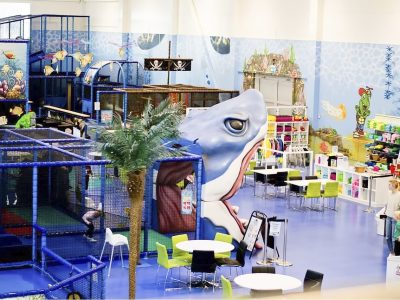En färgglad inomhuslekplats med ett marint tema, komplett med en stor hajslider, klätternät och en piratskeppssektion. I anslutning finns sittplatser med bord och stolar, samt en butik med leksaker och kläder.