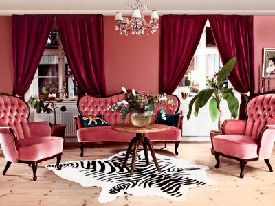 Ett elegant vardagsrum med två rosa fåtöljer, en rosa soffa, ett runt träbord och en zebrarandig matta. Rummet har röda sammetsgardiner framför två fönster, och en kristallkrona hänger från taket. I bakgrunden syns rosa väggar och ett grönt träd i en kruka.