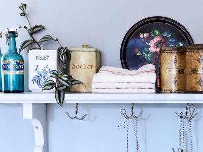 En vägghylla med vintage köksartiklar, inklusive två blå flaskor, en vit burk märkt "FRUKT", en gul burk märkt "Socker", gamla plåtburkar och en dekorativ bricka med blommor. En grön växt hänger ner från en av burkarna, och en stapel med vikta handdukar ligger mellan burkarna. Krokar hänger under hyllan.