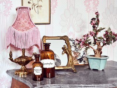 En antik skrivbordslampa med en rosa fransig lampskärm, två bruna apoteksflaskor märkta "Krumelurpiller" och "Kvickrotpulver Medicin", en guldram och en liten bonsai-liknande träd med rosa blommor, allt placerat på en marmorskiva. Bakom finns en ljusrosa tapet med blommönster.