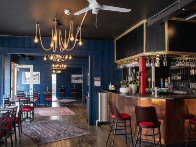 En restaurang med mörkblå väggar och tak. I förgrunden till höger finns en kopparfärgad bar med barstolar. Till vänster syns ett matrum med flera bord dukade med mörka dukar och röda stolar. Hela lokalen har trägolv och taket pryds av en fläkt samt unika taklampor gjorda av rep med synliga glödlampor.