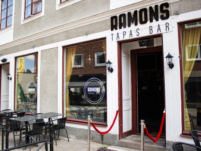 Framsidan av "Ramons Tapas Bar" i Vimmerby, med stora fönster, en entré med röda rep och utomhusbord med svarta stolar.