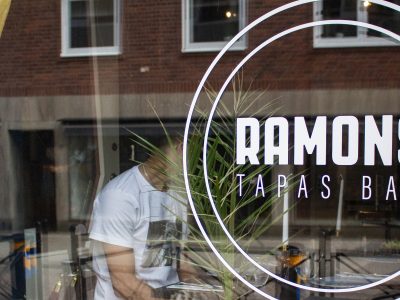 En närbild på fönstret till "Ramons Tapas Bar" i Vimmerby, med deras logotyp tydligt synlig och en reflektion av en person i en vit t-shirt som syns genom glaset.