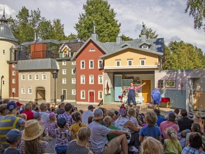 En stor publik, bestående av både barn och vuxna, tittar på en teaterföreställning utomhus. Scenen föreställer en stadsmiljö med färgglada byggnader, och skådespelare uppträder framför en interiörkuliss.