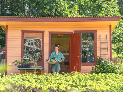 En leende kvinna håller en korg med grönsaker och står i dörröppningen till en orange byggnad märkt "Gårdsbutik Korka Kvarn". Framför byggnaden finns ett fält med gröna växter under en solig himmel.