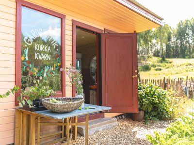 En ljusorange gårdsbutik med röda fönsterkarmar och dörr, där texten "Gårdsbutik Korka Kvarn" är målad på fönstret. Utanför står ett bord med en korg fylld med grönsaker och blommor. En öppen dörr leder in till butiken, och i bakgrunden syns skog och fält.