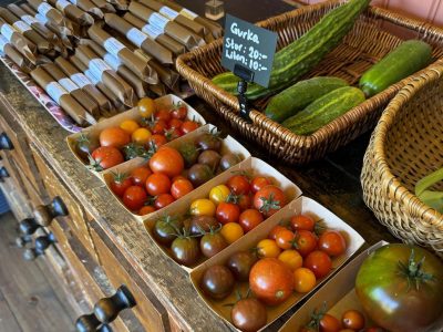 Ett bord med nyplockade grönsaker till salu. Bland annat syns lådor med blandade körsbärstomater i olika färger, korgar med gurkor och en skylt som anger priset för gurka: "Stor: 20:- Liten: 10:-". I bakgrunden finns även förpackade varor.