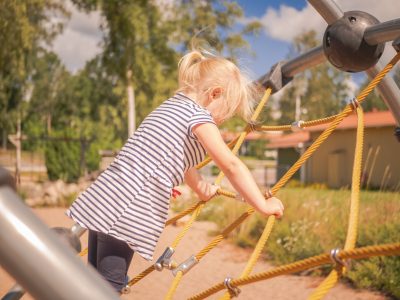 En blond flicka i en randig t-shirt klättrar på ett repnät på en lekplats utomhus.