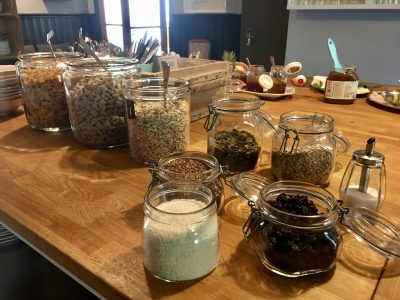 En närbild av en träbänk med olika torra ingredienser som müsli, frön och flingor i glasburkar, för en frukostbuffé.