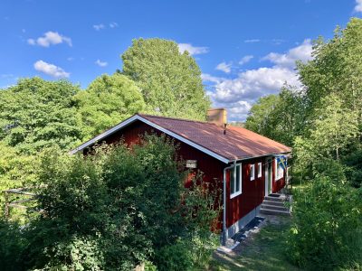 En charmig, röd stuga med vita knutar och ett rött tak, delvis dold av frodig grönska, med en svensk flagga hängande vid ingången.