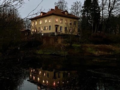 Ett upplyst gult hus med rött tak, omgivet av träd, reflekteras i en mörk damm vid skymning.