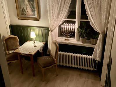 Ett mysigt rum i Vimmerby med mörkgröna paneler, ljusa väggar, ett litet bord med två stolar, och ett fönster med vita spetsgardiner och en adventsljusstake. En kristallkrona hänger från taket.