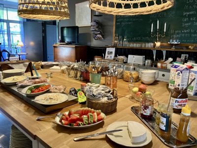 En frukostbuffé uppdukad på ett långt träbord i Vimmerby, med tallrikar fyllda med frukt, grönsaker, pålägg och flingor i glasburkar. I bakgrunden syns en svart griffeltavla med text, och två korglampor hänger över bordet.