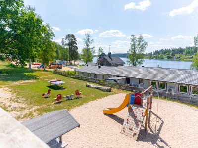En flygbild över Vimmerby campingplats vid en sjö, med en sandig lekplats med en rutschkana i förgrunden, omgiven av gräsytor och träd. Fler byggnader och båtar syns vid strandlinjen i bakgrunden.