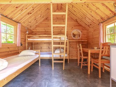 En rustik stuga med väggar och tak i ljust trä, med en våningssäng och en enkelsäng. Ett träbord med stolar står i mitten, och fönster med rödrutiga gardiner syns på vardera sida.