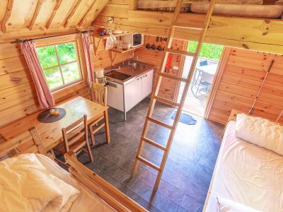 En bild tagen uppifrån inuti en liten trästuga med en stege som leder till ett loft. Stugan har ett pentry, en matplats med ett träbord och två stolar, samt två sängar. Ett fönster och en dörr med utsikt över grönska syns också.