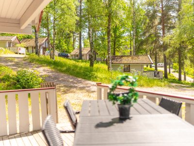 En veranda med ett utomhusbord och stolar, som vetter mot en grusväg med flera stugor och träd i bakgrunden.
