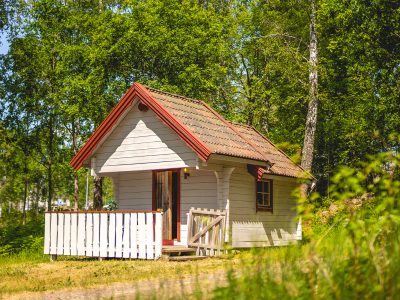 En liten, charmig vit stuga med rött tak och en veranda, omgiven av grönskande träd i Vimmerby.