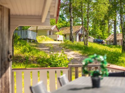 En veranda med ett utomhusbord och stolar, som vetter mot en grusväg med flera stugor och träd i bakgrunden.