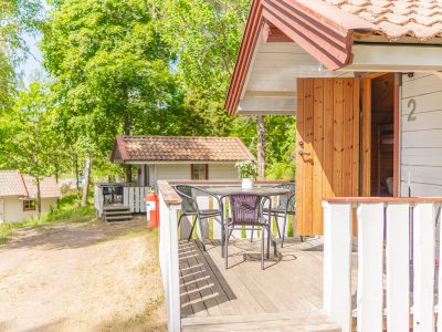 En träveranda med utemöbler och en öppen dörr till en stuga märkt med "2". I bakgrunden syns fler stugor och grönskande träd.
