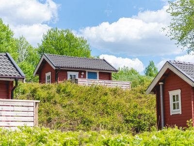 Tre röda stugor med mörka tak, omgivna av frodig grönska under en klarblå himmel med vita moln i Vimmerby.