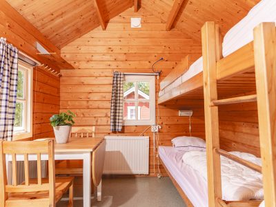 En trästuga i Vimmerby med en våningssäng, en enkelsäng, ett matbord och stolar. Fönster med rutiga gardiner släpper in ljus.