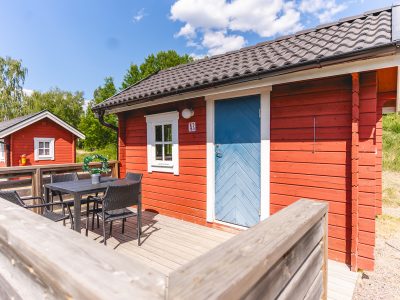 En röd stuga med blå dörr och mörkt tak, en träaltan med utemöbler, och en annan röd stuga i bakgrunden under en blå himmel med vita moln i Vimmerby.