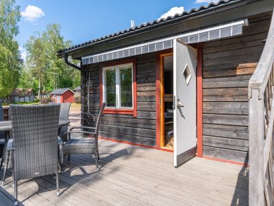 En mörk trähus med en träaltan och utemöbler. En öppen dörr leder in i huset, och ett fönster med röda foder syns intill. I bakgrunden syns fler stugor och träd under en blå himmel.