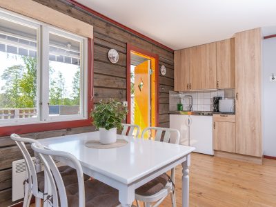 Ett rustikt inrett rum med timmerpanelväggar och ett ljust trägolv. Ett vitt matbord med stolar står i förgrunden, bredvid stora fönster som vetter mot exteriören. En öppen orange dörr leder ut. I bakgrunden syns ett litet kök med ljusa träskåp och vitvaror.