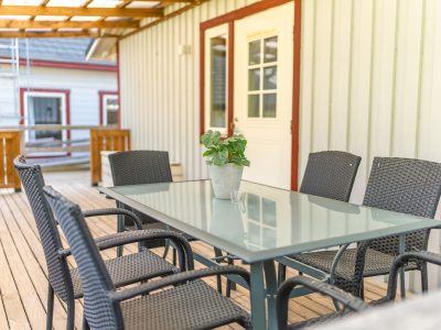 En träterrass med ett glasbord och mörkgrå stolar, med en krukväxt i mitten. En vit dörr med röda foder syns i bakgrunden.