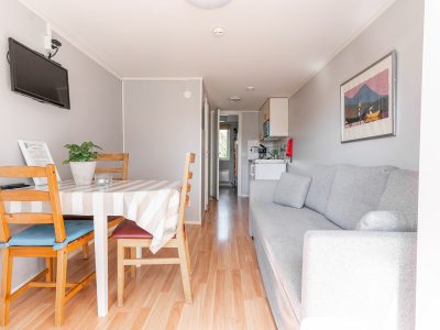 En liten stuga i Vimmerby med en öppen planlösning som visar ett vardagsrum med en grå soffa och en TV, och en matplats med ett randigt dukat bord. I bakgrunden syns ett litet kök.