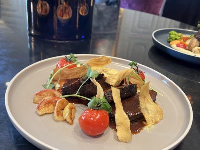En tallrik med en elegant upplagd maträtt, troligen långkokt kött med en mörk sås, garnerad med körsbärstomater, friterade chips och örter, placerad på ett mörkt bord med en stor vas i bakgrunden.