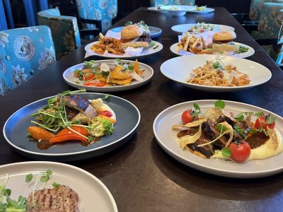 Ett långt matbord i en restaurang, täckt med en mängd olika rätter, inklusive biff, fisk, hamburgare och pasta, allt elegant upplagt. Runt bordet står stolar med blåmönstrade tyger.
