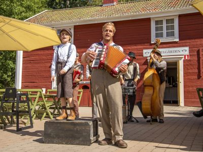 En utomhusscen framför en röd byggnad med flera musiker som spelar. Paradis Oskar spelar dragspel i förgrunden, bredvid honom står Rasmus som sjunger, och bakom dem finns en kontrabasist och en trummis. En gul parasoll skuggar delar av området.