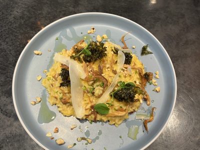 En tallrik med krämig risotto, toppad med rostade grönsaker, hyvlade grönsaker, hackade nötter och örter, serverad på ett mörkt bord.