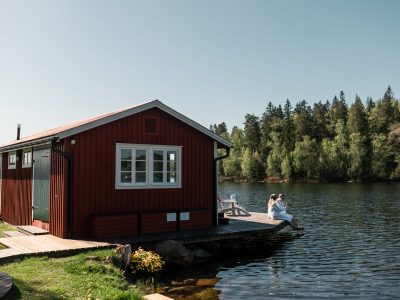 En röd stuga vid vattnet med en brygga där två personer sitter och tittar ut över sjön, omgiven av trädklädda stränder.