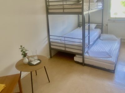 Ett enkelt sovrum med en våningssäng, en utdragbar säng och ett litet bord med en vas.