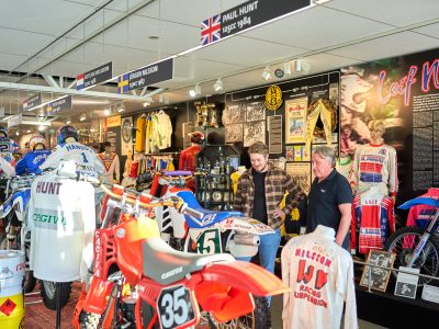 Två män tittar på motorcyklar och tävlingsutrustning i ett museum tillägnat motorsport. Displayerna inkluderar gamla tävlingscyklar, tröjor och informationstavlor om kända förare som Leif Nilsson.