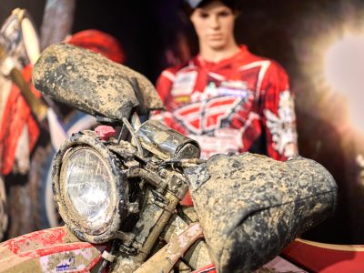 En lerig motocrosscykel i förgrunden, med en realistisk skyltdocka i röd motocrossutrustning i bakgrunden.